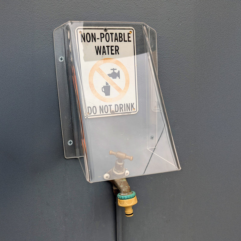 A clear plastic water tap cover mounted on a dark gray wall, with a white sign that reads 'NON-POTABLE WATER' and 'DO NOT DRINK' above a small faucet spout.