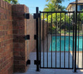 A black metal pool gate with vertical bars, installed next to a red brick wall overlooking a swimming pool and backyard garden. A clear acrylic panel fills a gap between the metal pool gate and brick wall.