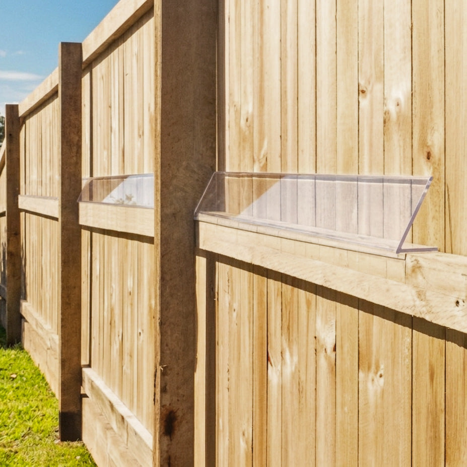 A clear plastic fence guard or cover mounted on a wooden garden fence, shown outdoors with grass and blue sky visible.