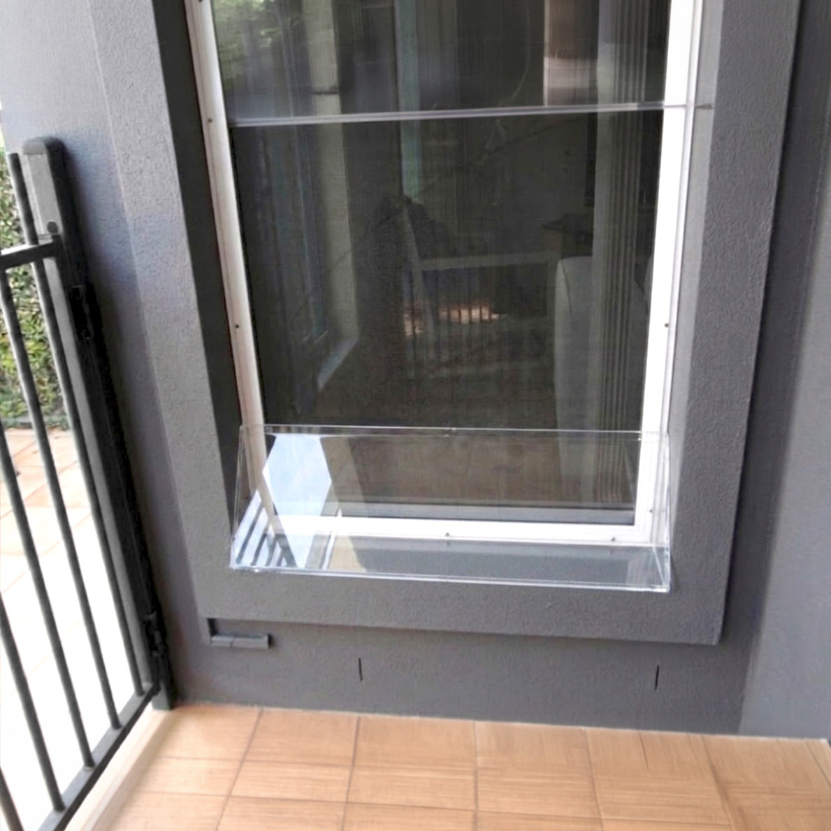 A clear plastic window screen door extension installed on a gray-framed window, shown on a tiled balcony next to a black metal railing.