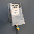 A clear plastic water tap cover mounted on a dark gray wall, with a white sign that reads 'NON-POTABLE WATER' and 'DO NOT DRINK' above a small faucet spout.
