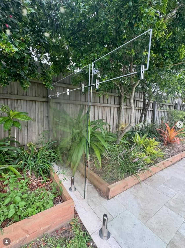 A glass privacy fence panel with metal clips holding clear acrylic panels on top to increase the fence height, in a backyard garden next to a paved walkway.