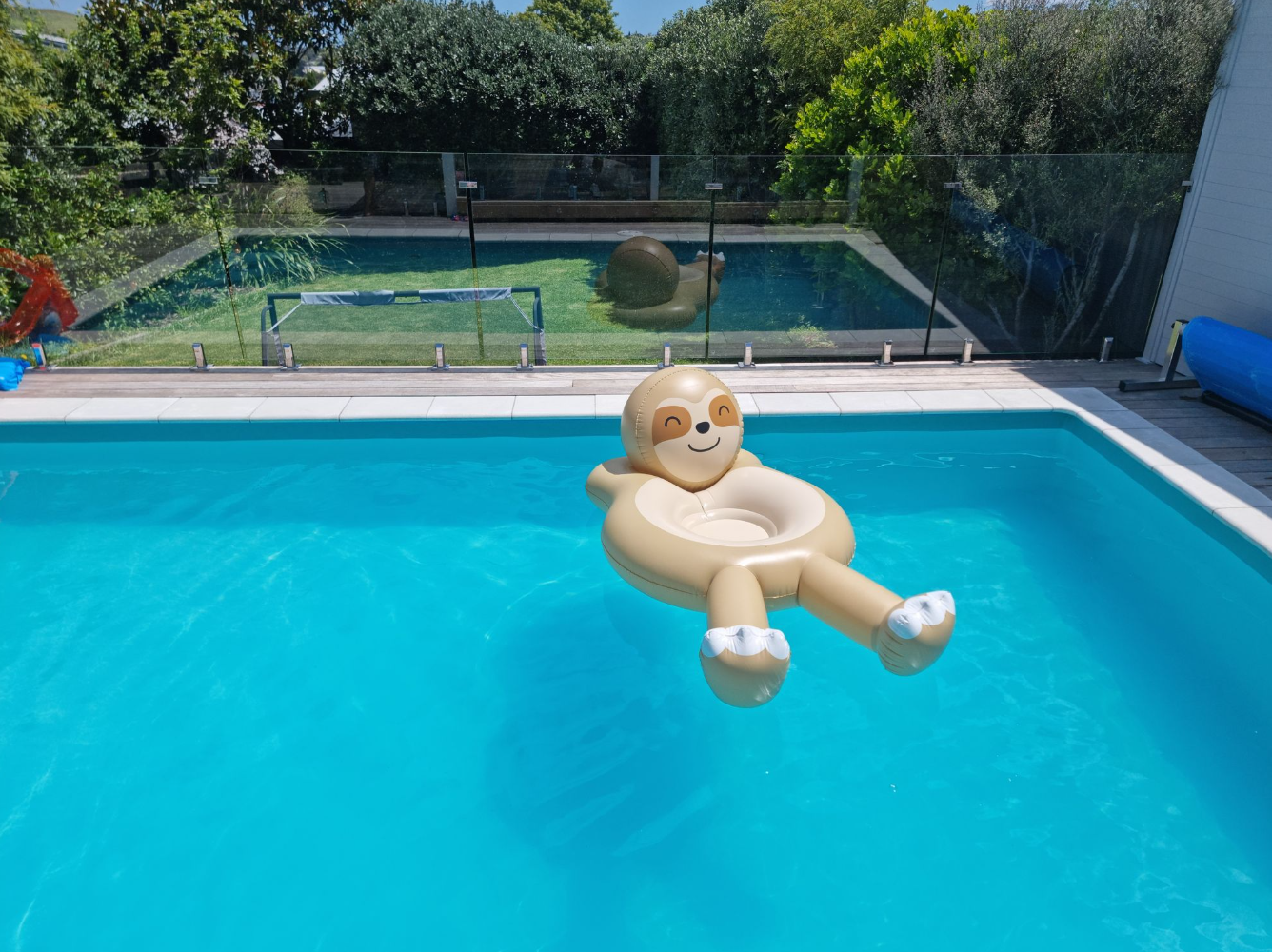 A beige sloth-shaped inflatable pool float with white feet floating in a bright blue swimming pool, with a backyard, trees, and another pool visible in the background.